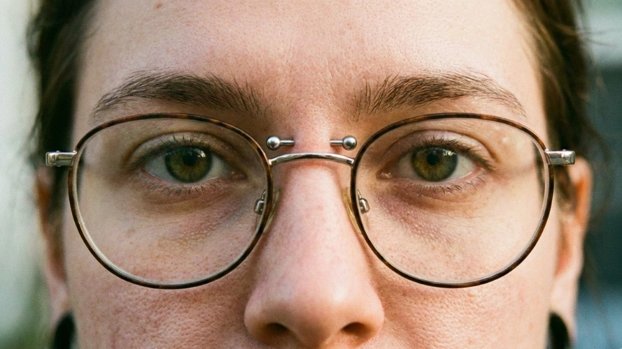 Gros plan sur un visage portant des lunettes rondes et un piercing "Bridge" horizontal sur l'arête du nez. Le piercing est orné de deux billes en titane.